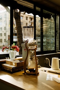 A wooden coffee bar setup with a cold brew coffee maker featuring a glass carafe and a wooden frame. Sunlight filters in through large windows, casting shadows on the wooden counter. Beside the coffee maker, there are red and white flowers in a vase, stirrers, and a box for napkins. A kettle and coffee-related papers are also visible.