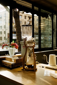 A wooden coffee bar setup with a cold brew coffee maker featuring a glass carafe and a wooden frame. Sunlight filters in through large windows, casting shadows on the wooden counter. Beside the coffee maker, there are red and white flowers in a vase, stirrers, and a box for napkins. A kettle and coffee-related papers are also visible.