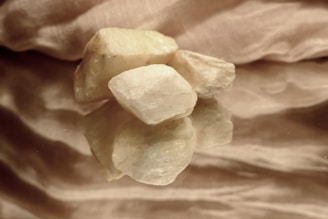 Several pale, irregularly shaped stones are placed on a reflective surface, with a textured fabric backdrop. The lighting is soft, highlighting the rough texture and natural color variations of the stones, creating an elegant and serene composition.