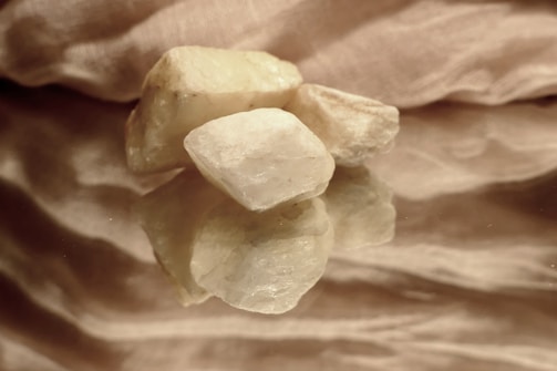 Several pale, irregularly shaped stones are placed on a reflective surface, with a textured fabric backdrop. The lighting is soft, highlighting the rough texture and natural color variations of the stones, creating an elegant and serene composition.