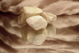 Several pale, irregularly shaped stones are placed on a reflective surface, with a textured fabric backdrop. The lighting is soft, highlighting the rough texture and natural color variations of the stones, creating an elegant and serene composition.
