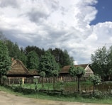 A rural landscape featuring traditional thatched-roof cottages surrounded by lush green trees. A rustic wooden fence encloses the area, with a small pond reflecting the cloudy sky above. The scene has a peaceful, countryside vibe.