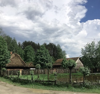 A rural landscape featuring traditional thatched-roof cottages surrounded by lush green trees. A rustic wooden fence encloses the area, with a small pond reflecting the cloudy sky above. The scene has a peaceful, countryside vibe.