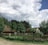 A rural landscape featuring traditional thatched-roof cottages surrounded by lush green trees. A rustic wooden fence encloses the area, with a small pond reflecting the cloudy sky above. The scene has a peaceful, countryside vibe.