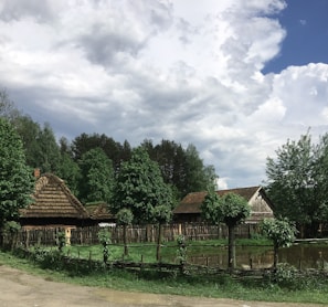 A rural landscape featuring traditional thatched-roof cottages surrounded by lush green trees. A rustic wooden fence encloses the area, with a small pond reflecting the cloudy sky above. The scene has a peaceful, countryside vibe.