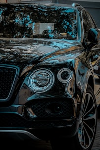 A luxury SUV with a glossy black finish, reflecting surrounding trees and sky. The vehicle features distinctive round headlights and a sleek grille.