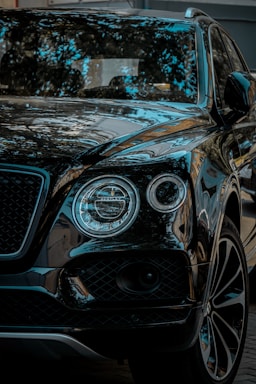 A luxury SUV with a glossy black finish, reflecting surrounding trees and sky. The vehicle features distinctive round headlights and a sleek grille.