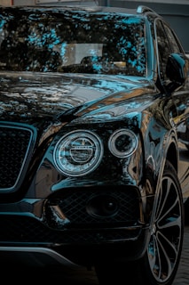 A luxury SUV with a glossy black finish, reflecting surrounding trees and sky. The vehicle features distinctive round headlights and a sleek grille.
