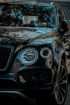 A luxury SUV with a glossy black finish, reflecting surrounding trees and sky. The vehicle features distinctive round headlights and a sleek grille.