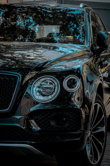 A luxury SUV with a glossy black finish, reflecting surrounding trees and sky. The vehicle features distinctive round headlights and a sleek grille.