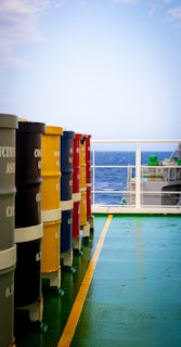 Brightly colored barrels are lined up on a ship's deck, overlooking the ocean under a clear sky. The deck has a green and yellow painted surface leading towards a railing that shows the expanse of water and part of the ship's structure.