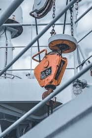 A close-up view of a metallic industrial hook system hanging from chains. The hook is painted orange and marked with '0.9t', indicating its load capacity. The background reveals the structure of a ship, with diagonal beams and a cloudy sky.