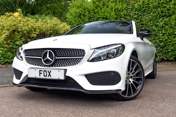 A sleek white Mercedes-Benz car is parked on a concrete surface, surrounded by lush green hedges and trees. The car has a distinctive grille with a prominent Mercedes emblem and a license plate labeled 'FOX'. The sleek design includes modern headlights and large alloy wheels, conveying luxury and performance.