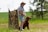 An older man is standing outdoors on a grassy field with a large brown dog sitting beside him. The man is wearing a sun hat, sunglasses, a checkered shirt, denim shorts, and sneakers. Behind them is a camouflage-patterned fabric screen and a landscape of green grass and trees under a partly cloudy sky.