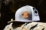 A trucker cap featuring a gray and white design with a circular patch labeled 'DERBY SAW & GENERAL STORE EST. 2015'. There is a tag attached that reads 'CAMBRIDGE'. The cap is placed on a rugged surface with tire-like treads in the background, suggesting an outdoor, rustic setting.