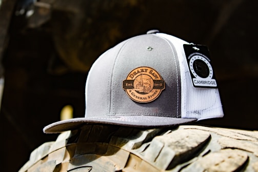 A trucker cap featuring a gray and white design with a circular patch labeled 'DERBY SAW & GENERAL STORE EST. 2015'. There is a tag attached that reads 'CAMBRIDGE'. The cap is placed on a rugged surface with tire-like treads in the background, suggesting an outdoor, rustic setting.