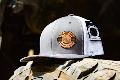 A trucker cap featuring a gray and white design with a circular patch labeled 'DERBY SAW & GENERAL STORE EST. 2015'. There is a tag attached that reads 'CAMBRIDGE'. The cap is placed on a rugged surface with tire-like treads in the background, suggesting an outdoor, rustic setting.