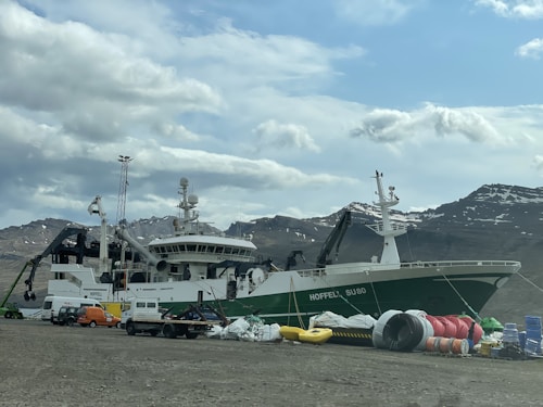 A large fishing vessel is docked near a rocky shore, with a range of equipment and several vehicles surrounding it. The ship, named HOFFELL, is painted green and white, and the background features rugged mountains with patches of snow under a cloudy sky.
