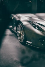 A sleek, high-performance sports car with a metallic finish is partially visible in the image. The focus is on the front right side, showing an aerodynamic design with polished alloy wheels and a prominent headlight. The logo on the car's side is noticeable, and the vehicle is parked indoors, reflecting ambient light.