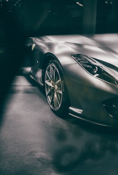 A sleek, high-performance sports car with a metallic finish is partially visible in the image. The focus is on the front right side, showing an aerodynamic design with polished alloy wheels and a prominent headlight. The logo on the car's side is noticeable, and the vehicle is parked indoors, reflecting ambient light.