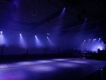 A dimly lit indoor space with blue and purple lighting against the ceiling and walls. A series of spotlights cast focused beams across the large empty floor area. The setting appears to be spacious with a raised stage or DJ booth visible in the background on the right side, slightly obscured by fog or smoke effects. The environment suggests a nightclub or concert venue setup, preparing for an event.