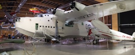 A large, historic aircraft is displayed indoors within a museum setting. The aircraft is primarily white with black propellers and is accompanied by several other smaller aircrafts in the background. The space is well-lit and spacious, showcasing the vintage aircraft with detailed attention, including maintenance access stairs and ropes to keep visitors at a distance.