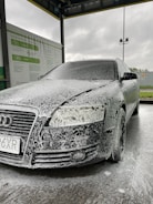 A car is covered in soapy foam at a car wash station. The vehicle appears to be dark-colored, possibly black, with a layer of white foam applied over it, giving a speckled appearance. The car is parked under a shelter with a metal roof, and a sign with instructions or information is visible on the side. The background shows a cloudy sky, suggesting an overcast day.