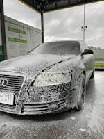 A car is covered in soapy foam at a car wash station. The vehicle appears to be dark-colored, possibly black, with a layer of white foam applied over it, giving a speckled appearance. The car is parked under a shelter with a metal roof, and a sign with instructions or information is visible on the side. The background shows a cloudy sky, suggesting an overcast day.