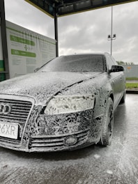 A car is covered in soapy foam at a car wash station. The vehicle appears to be dark-colored, possibly black, with a layer of white foam applied over it, giving a speckled appearance. The car is parked under a shelter with a metal roof, and a sign with instructions or information is visible on the side. The background shows a cloudy sky, suggesting an overcast day.