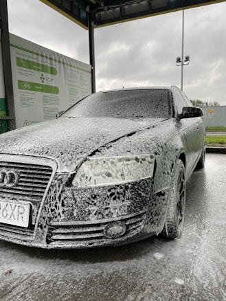 A car is covered in soapy foam at a car wash station. The vehicle appears to be dark-colored, possibly black, with a layer of white foam applied over it, giving a speckled appearance. The car is parked under a shelter with a metal roof, and a sign with instructions or information is visible on the side. The background shows a cloudy sky, suggesting an overcast day.