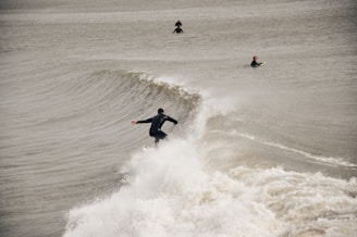 Three surfers are in the ocean, with one riding a wave while the other two are waiting in the water. The waves are strong, and the water appears choppy. All three are wearing dark wetsuits.