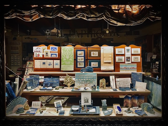 A display window showcasing an assortment of paper products and stationery, including notebooks, postcards, framed art prints, and decorative paper. The items are organized on several shelves, with an emphasis on blue and green tones, and feature intricate designs and patterns that suggest a focus on fine craftsmanship and artistic detail.
