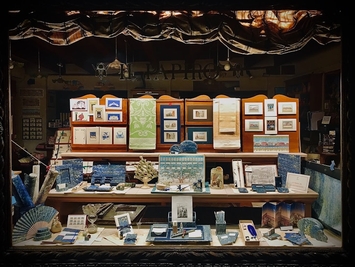 A display window showcasing an assortment of paper products and stationery, including notebooks, postcards, framed art prints, and decorative paper. The items are organized on several shelves, with an emphasis on blue and green tones, and feature intricate designs and patterns that suggest a focus on fine craftsmanship and artistic detail.