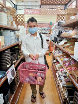 A person wearing a mask stands in a store aisle holding a pink shopping basket. The store shelves are stocked with various kitchenware items and other household goods. The person is wearing a white shirt, brown pants, and flip-flops.