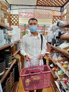 A person wearing a blue face mask and white shirt is holding a pink shopping basket in a store aisle. The shelves are filled with various home goods such as baskets, pots, and kitchenware.