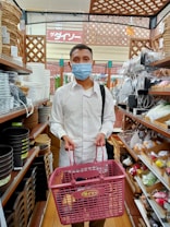 A person wearing a blue face mask and white shirt is holding a pink shopping basket in a store aisle. The shelves are filled with various home goods such as baskets, pots, and kitchenware.