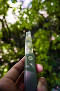 A hand holding a pocket knife with a decorative blade, featuring intricate patterns. The background is a blur of green foliage, suggesting a natural outdoor setting.