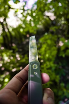 A hand holding a pocket knife with a decorative blade, featuring intricate patterns. The background is a blur of green foliage, suggesting a natural outdoor setting.