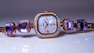 A luxurious women's wristwatch with a square, mother-of-pearl dial, framed by a ring of diamonds. The strap features large, square purple gemstones, creating an elegant and sophisticated look. The watch hands and markers are minimalistic, with a brand name visible on the dial.