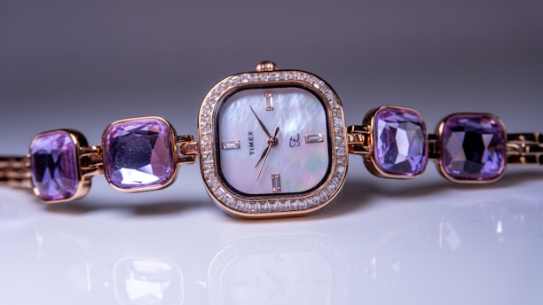 A luxurious women's wristwatch with a square, mother-of-pearl dial, framed by a ring of diamonds. The strap features large, square purple gemstones, creating an elegant and sophisticated look. The watch hands and markers are minimalistic, with a brand name visible on the dial.