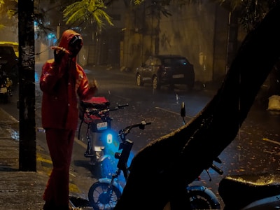 A person in a red raincoat stands on a wet street under low light. They are close to a bicycle, which has a glowing light, and the street is lined with parked cars and trees. Raindrops can be seen falling, creating a moody and rainy atmosphere.