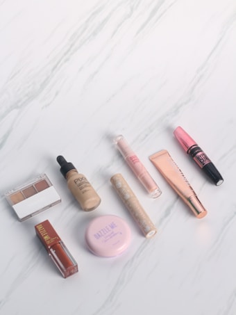 A collection of various makeup products arranged on a light marbled surface, including lip gloss, foundation, eyeshadow, and blush, with sleek and colorful packaging.