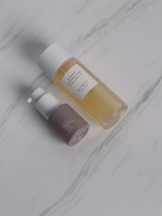 Two cosmetic bottles are placed on a light marble surface. One is a transparent bottle filled with a yellow liquid and has a white cap, labeled as botanical calendula oil. The other is a smaller pump bottle with a brown base and white top.