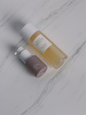 Two cosmetic bottles are placed on a light marble surface. One is a transparent bottle filled with a yellow liquid and has a white cap, labeled as botanical calendula oil. The other is a smaller pump bottle with a brown base and white top.