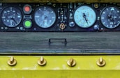 An industrial control panel features several large round dials with numbers and needle indicators. Adjacent are colored buttons, including bright red and green. The panel sits above a flat textured surface, and a row of rounded knobs is aligned at the bottom, all against a yellow background.