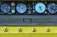 An industrial control panel features several large round dials with numbers and needle indicators. Adjacent are colored buttons, including bright red and green. The panel sits above a flat textured surface, and a row of rounded knobs is aligned at the bottom, all against a yellow background.