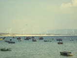 Several small fishing boats float on a calm sea with a distant shoreline featuring wind turbines. The water is a tranquil blue, and the sky is lightly overcast with clouds.