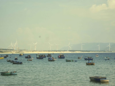Several small fishing boats float on a calm sea with a distant shoreline featuring wind turbines. The water is a tranquil blue, and the sky is lightly overcast with clouds.