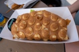 A bubble waffle with a golden brown appearance served in a white paper tray on a wooden table.