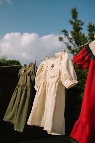 A line of children's clothes hanging on a clothesline outdoors. The dresses are in olive green, cream, and red colors, gently swaying in the breeze. The background features a clear blue sky with fluffy clouds and some green foliage.